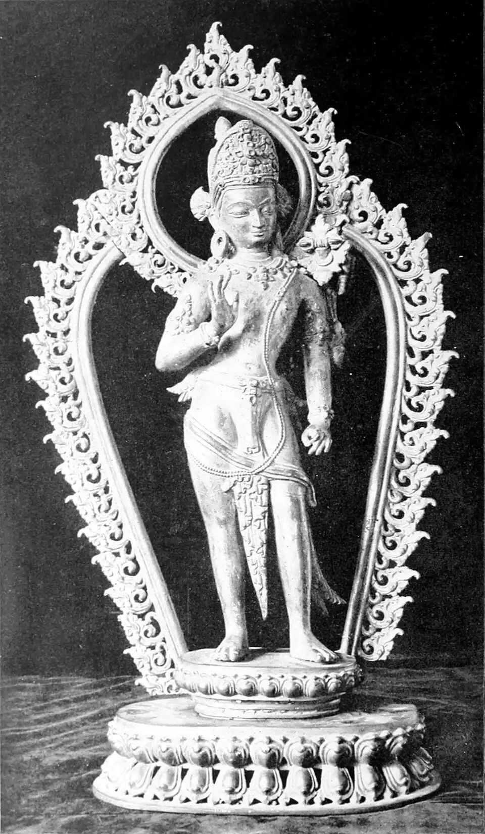 A black and white photograph of a figure standing on a round base, surrounded by an ornate arch. The right arm raised with palm forward at chest level; the left arm held by its side.