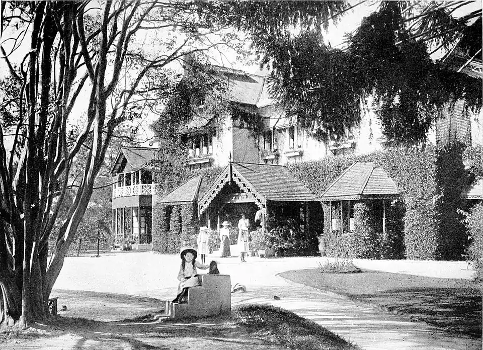 Black and white photograph of a large two storey house in the country. Four women stand in the background, by the front door. A girl sits on steps in the foreground, in the shade of a tree.